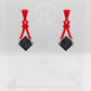 Image of a pair of acrylic earrings. The design is a thin elegant top that forks out to support a diamond shape dangling under it. They are Red for the top and a Black and Silver Glitter diamond. They are against a white background on a clear stand.