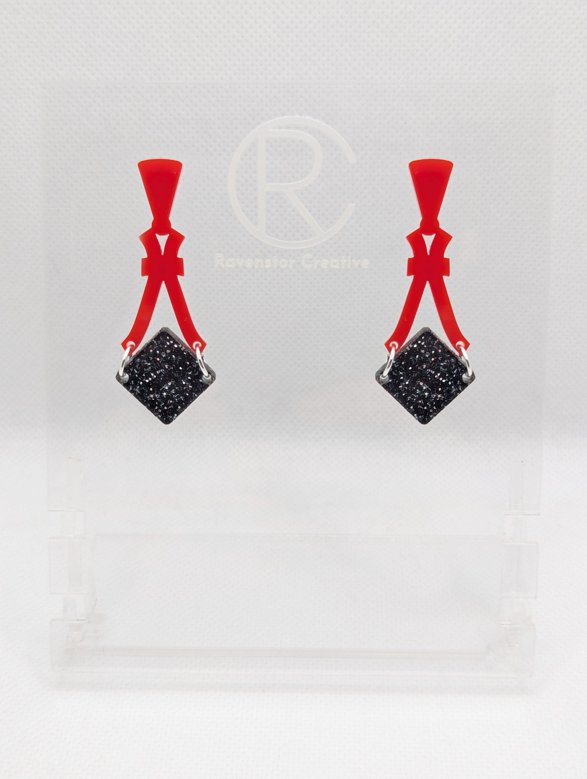 Image of a pair of acrylic earrings. The design is a thin elegant top that forks out to support a diamond shape dangling under it. They are Red for the top and a Black and Silver Glitter diamond. They are against a white background on a clear stand.