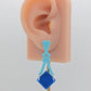 Image of a pair of acrylic earrings. The design is a thin elegant top that forks out to support a diamond shape dangling under it. They are Spearmint for the top and a Blue diamond. They are on an ear to show the size.