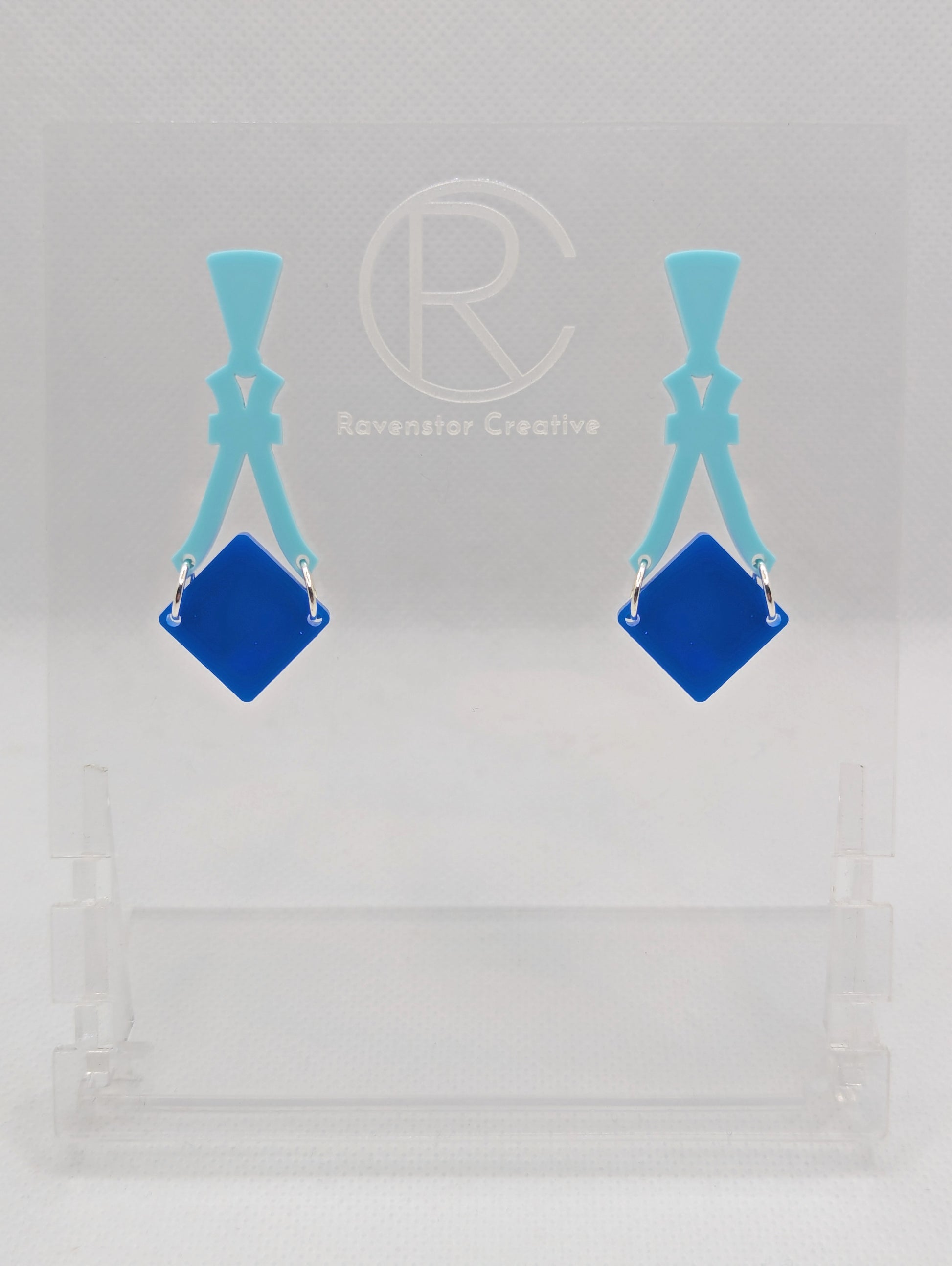 Image of a pair of acrylic earrings. The design is a thin elegant top that forks out to support a diamond shape dangling under it. They are Spearmint for the top and a Blue diamond. They are against a white background on a clear stand.