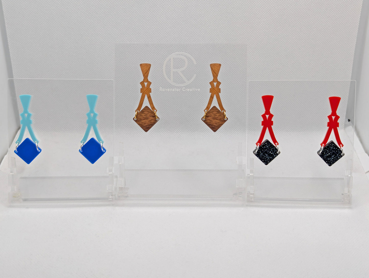 Image of three pairs of earrings. The design is a thin elegant top that forks out to support a diamond shape dangling under it. They are Spearmint and Blue, Red and Black Silver Acrylic and Cherry and Walnut Wood.