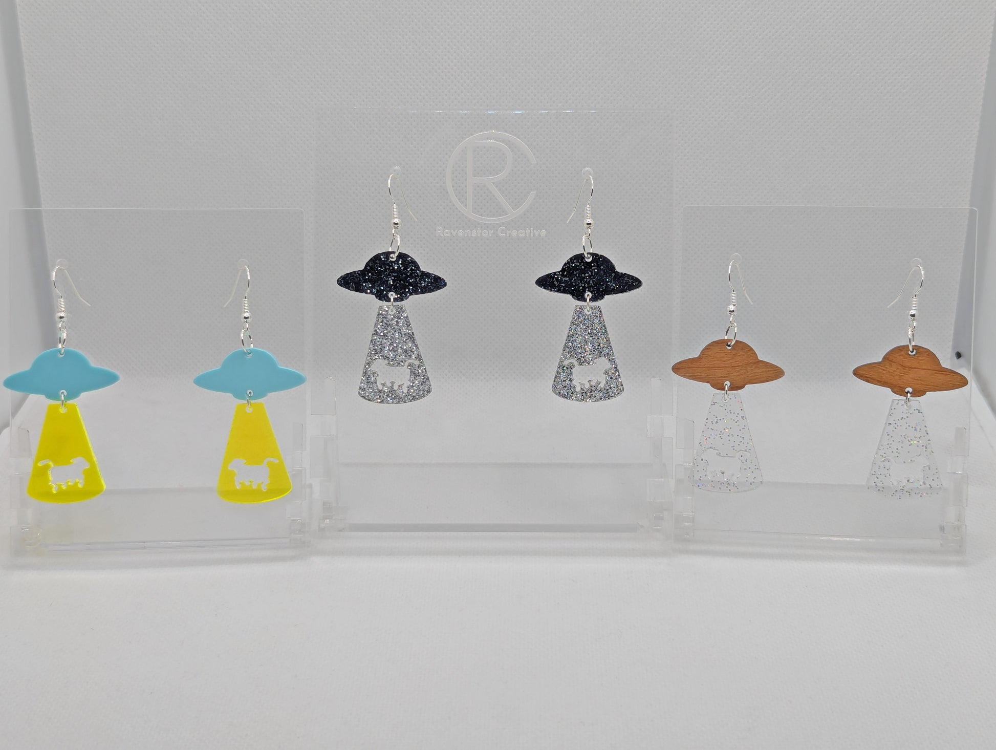 Three pairs of Flying Saucer earrings. They are two parts with a ship at the top and tractor beam underneath with the image of a cow cut out. The colours are Spearmint and Neon Orange, Black and Silver Glitter, Cherry Wood and clear sparkle.