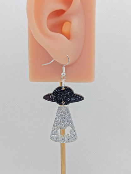 A Flying Saucer earring. They are a ship at the top and tractor beam underneath with the image of a cow cut out. The colours are Black and Silver glitter for the spaceship and Silver glitter for the tractor beam. It is on an ear to show the size.