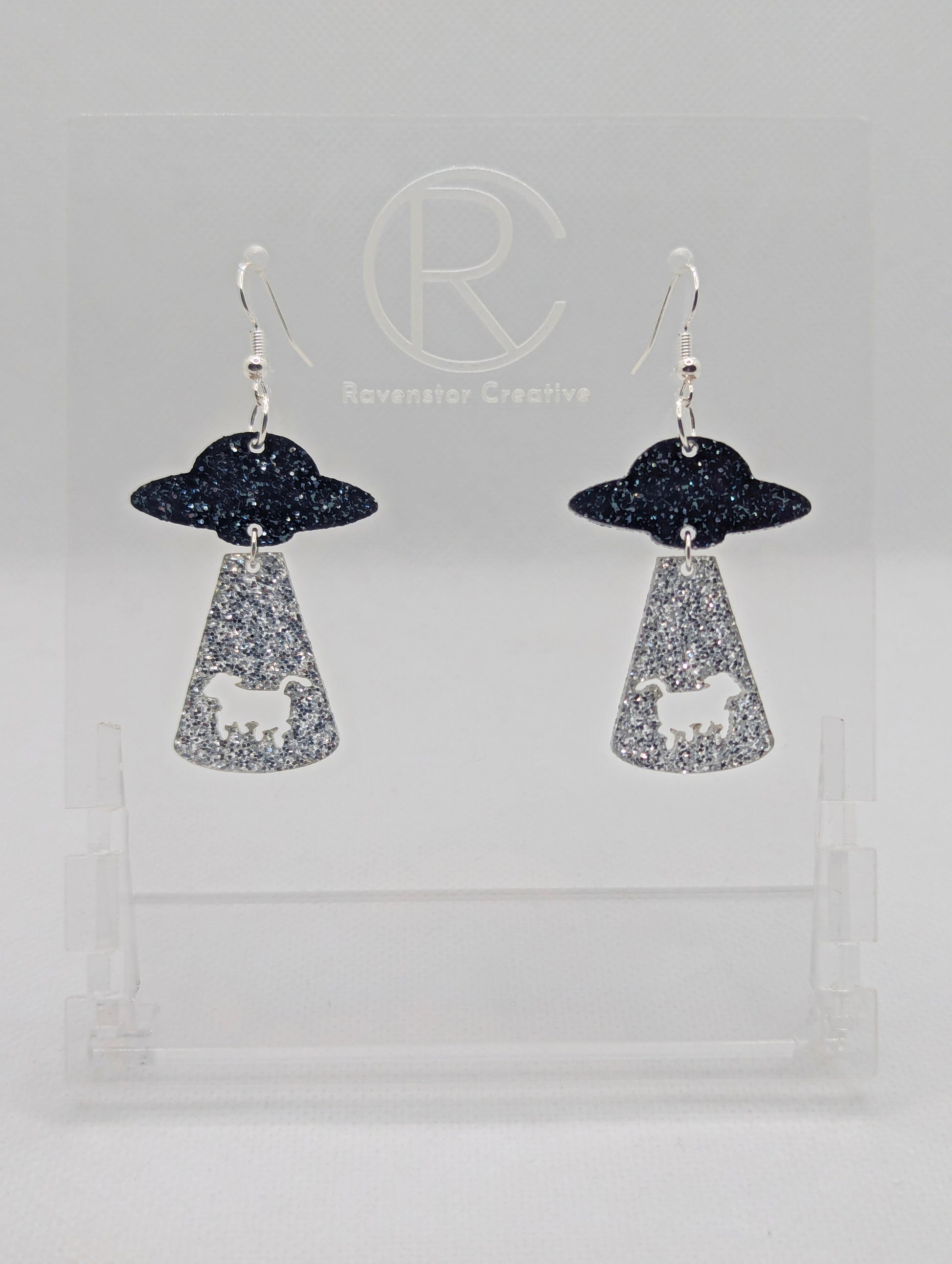 A pair of two part earrings with Silver ear wire hook fixings. The top part is a flying saucer and underneath there is a tractor beam with a cow cut out. The colours are Black and Silver Glitter for the ship and Silver Glitter for the tractor beam.