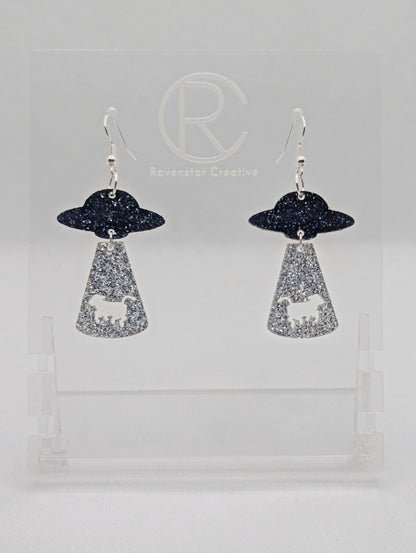 A pair of two part earrings with Silver ear wire hook fixings. The top part is a flying saucer and underneath there is a tractor beam with a cow cut out. The colours are Black and Silver Glitter for the ship and Silver Glitter for the tractor beam.