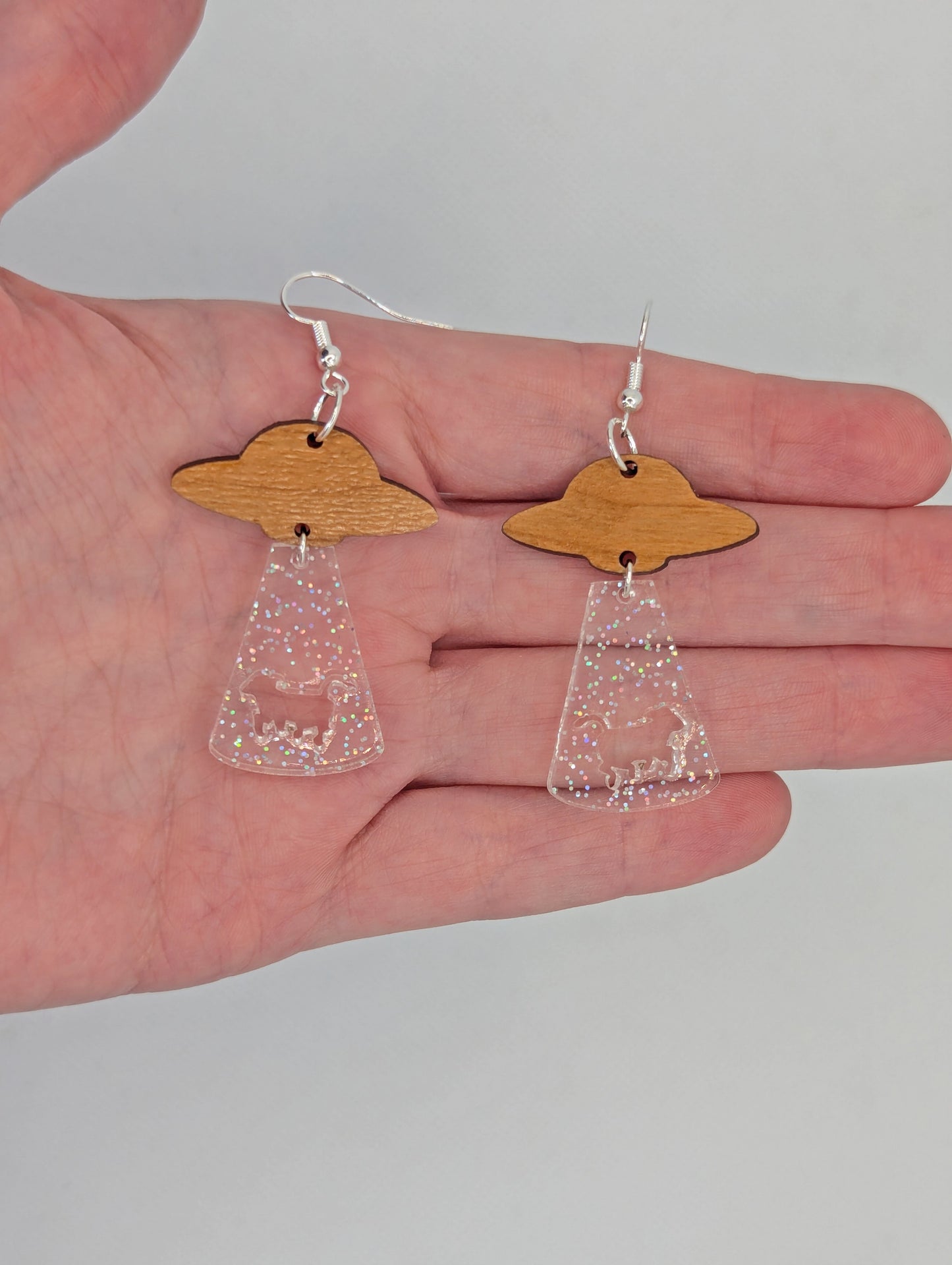 Flying Saucer earrings. They are a ship at the top and tractor beam underneath with the image of a cow cut out. They are against a hand to show the size of three finger widths in length and one and a half finger widths wide.