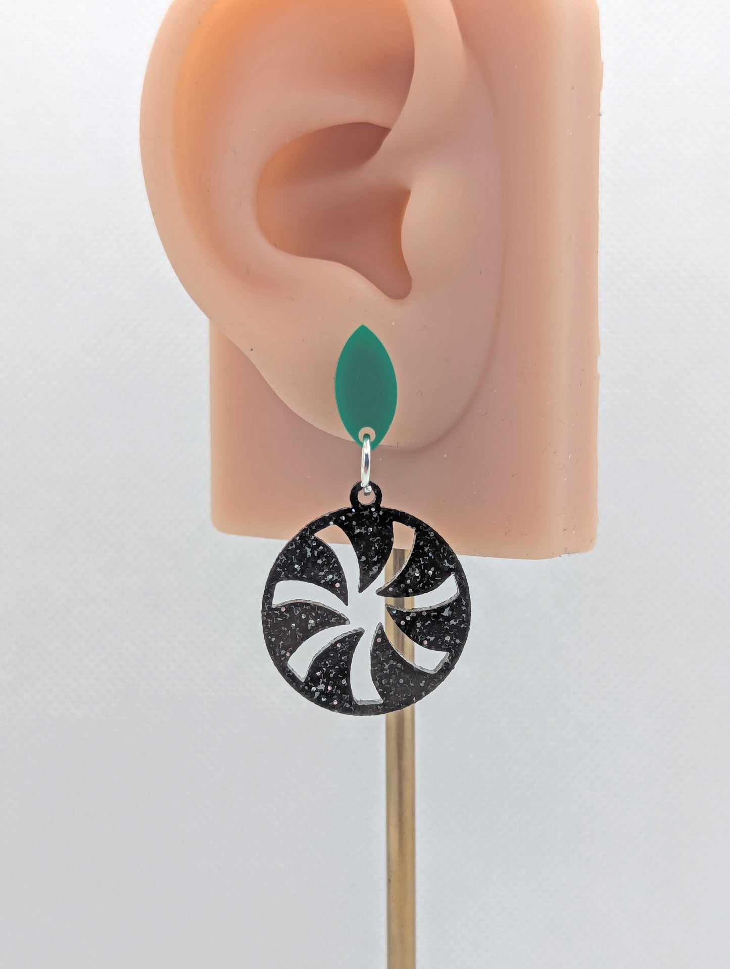 Acrylic stud earrings. They are a small diamond shape with a spiral cut out of a circle dangling underneath. The colours are Green and Black Silver and Glitter. They are on an ear to show the size.