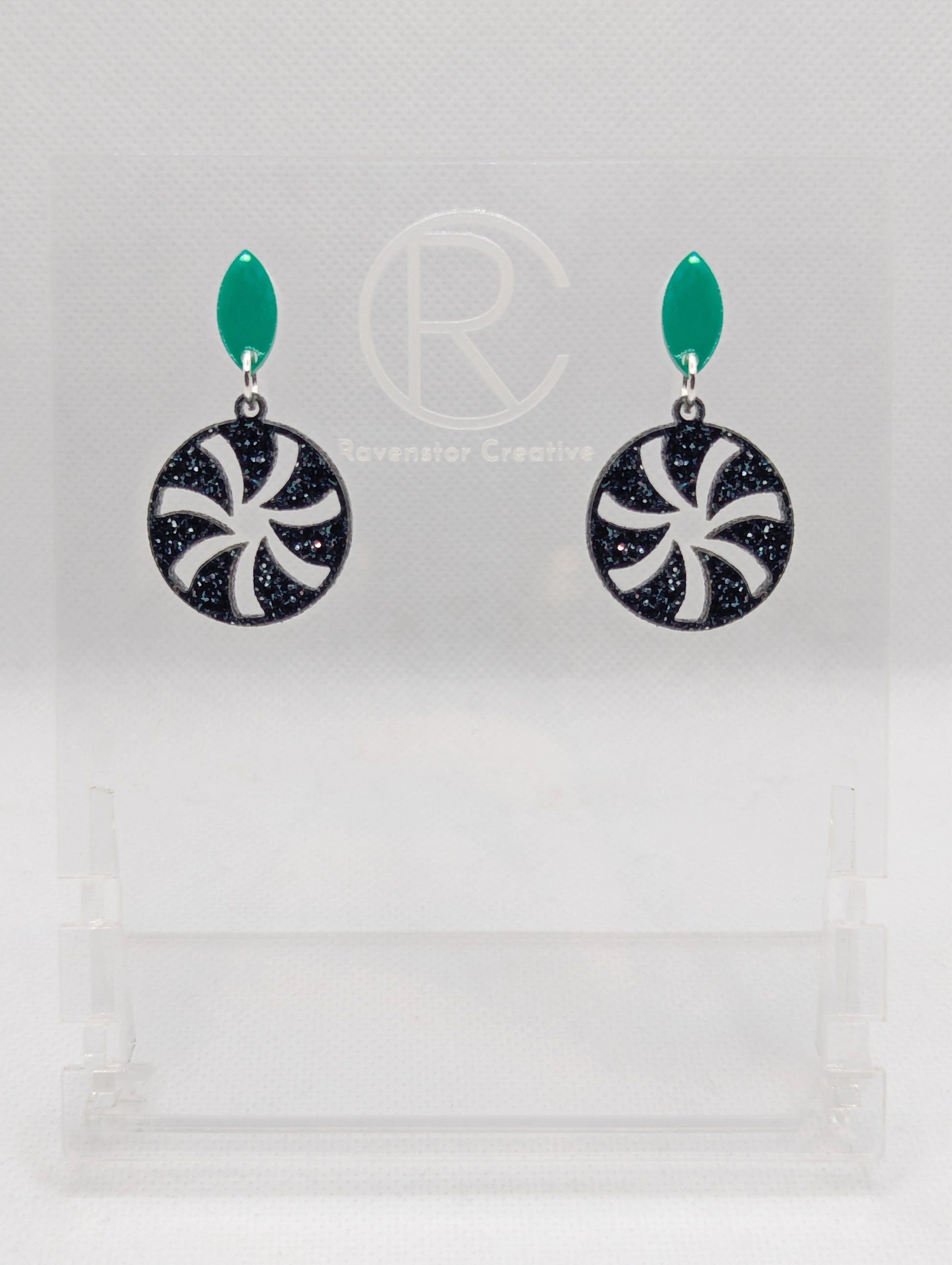 Acrylic stud earrings. They are a small diamond shape with a spiral cut out of a circle dangling underneath. The colours are Green and Black Silver and Glitter. They are on a clear stand with the Ravenstor Creative Logo against a white background.