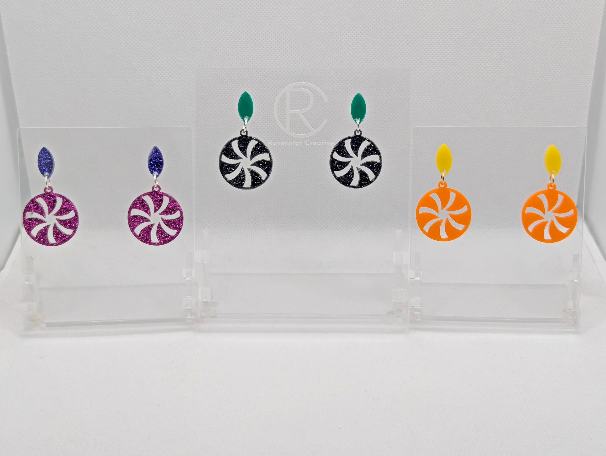 Three pairs of acrylic stud earrings. They are a small diamond shape with a spiral cut out of a circle dangling underneath. The colours are Purple and Pink Glitter. Green with Black and silver glitter. Yellow and orange.