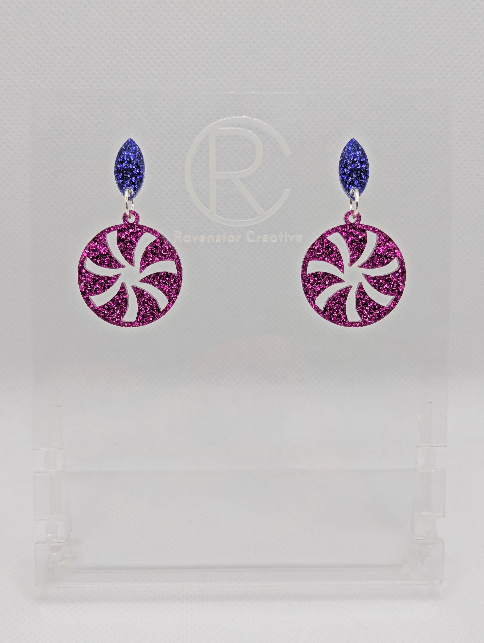 A pair of acrylic stud earrings. They are a small diamond shape with a spiral cut out of a circle dangling underneath. The colours are Purple and Pink Glitter. They are on a clear stand with the Ravenstor Creative Logo against a white background.