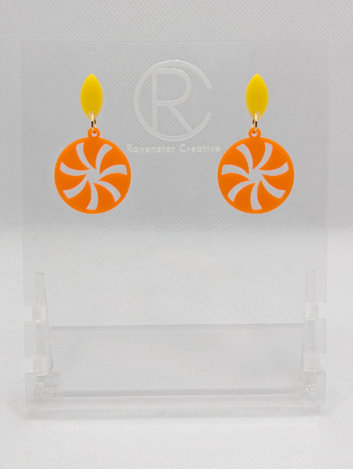 A pair of acrylic stud earrings. They are a small diamond shape with a spiral cut out of a circle dangling underneath. The colours are Yellow and Orange. They are on a clear stand with the Ravenstor Creative Logo against a white background.