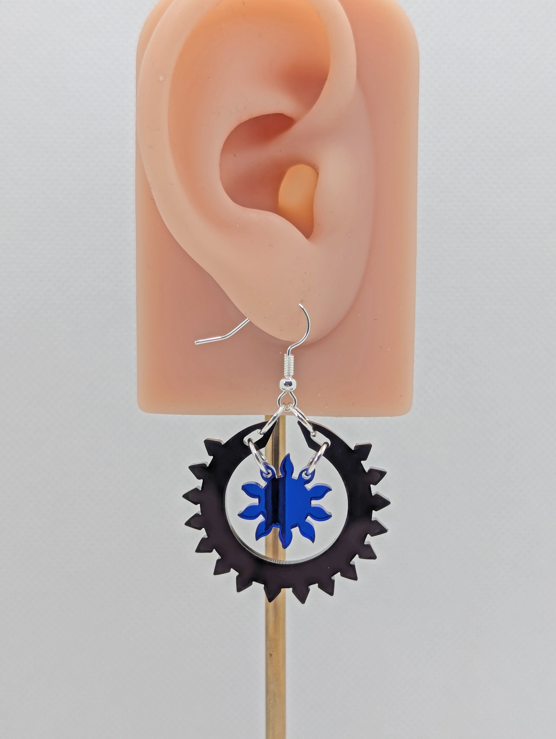 An image of a drop earring with a silver ear wire hook fixing, it is in two parts. The outer circle has points around it with a small sun shape in the middle. It is on an ear to show the size.