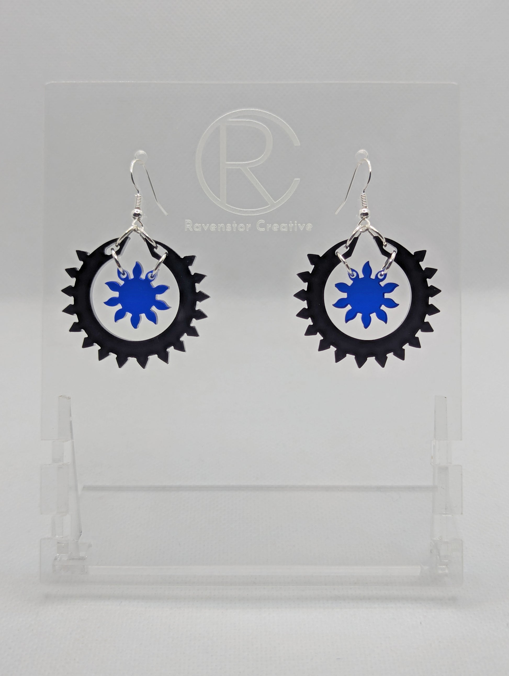 An image of a pair of drop earrings with silver ear wire hook fixings, each in two parts. The outer circle has points around it with a small sun shape in the middle. The colours are Black for the outer ring and Translucent Blue for the sun.