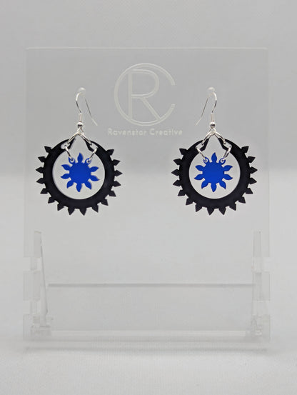 An image of a pair of drop earrings with silver ear wire hook fixings, each in two parts. The outer circle has points around it with a small sun shape in the middle. The colours are Black for the outer ring and Translucent Blue for the sun.