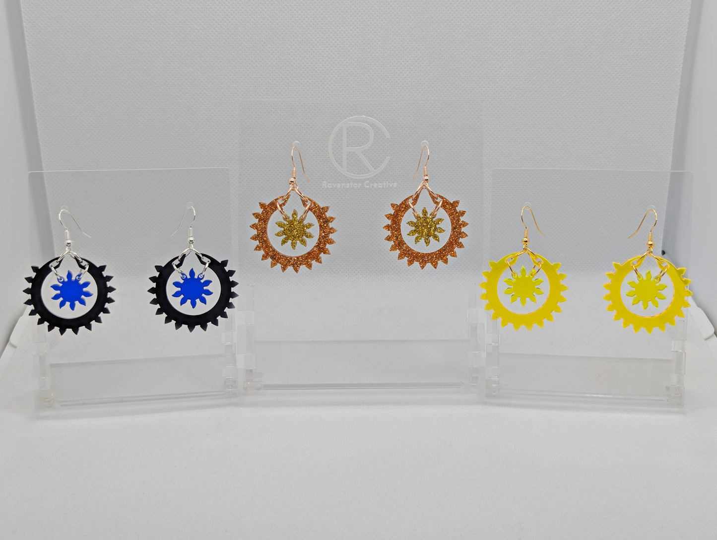 An image of three pairs of drop earrings, each in two parts. The outer circle has points around it with a small sun shape in the middle. The colours are Black and Translucent Blue, Copper Glitter and Gold Glitter, Neon Orange and Neon Yellow.