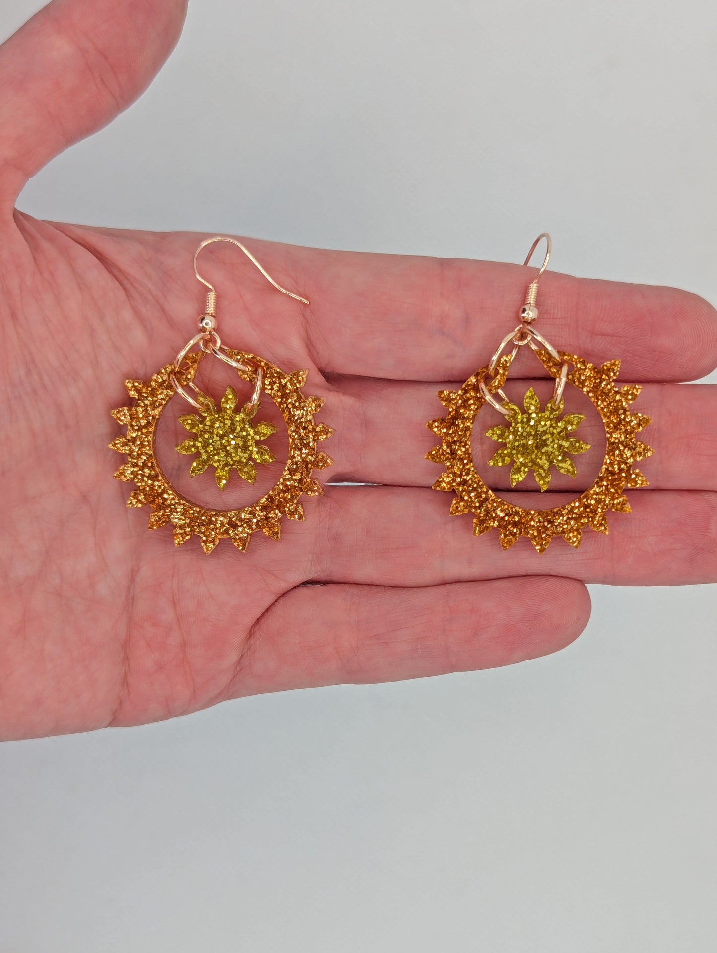 An image of a pair of drop earrings with Rose Gold ear wire hook fixings, each is in two parts. The outer circle has points around it with a small sun shape in the middle. It is on a hand to show the size of two finger widths in length and width.