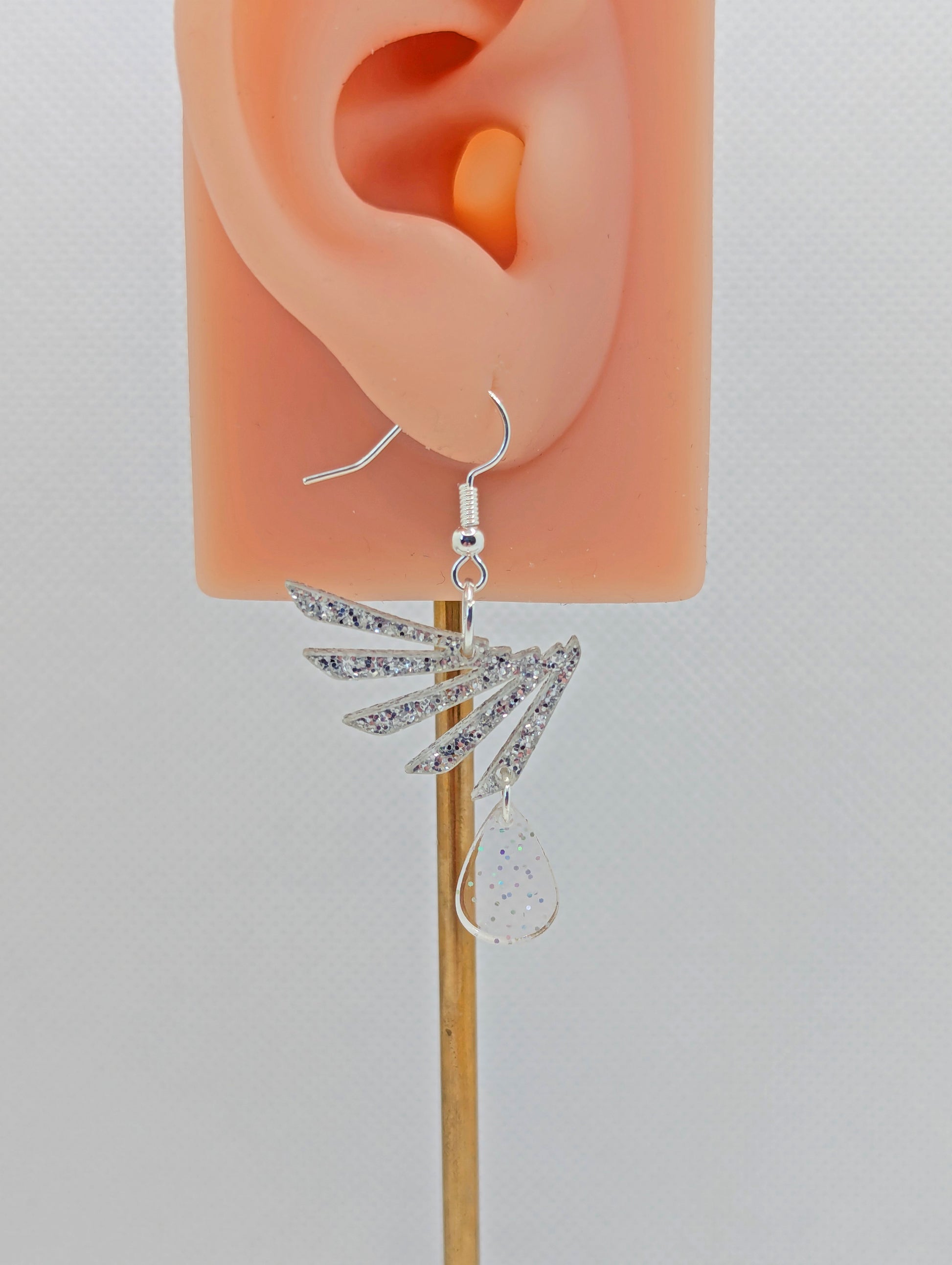 A two part drop earring with a Silver ear wire hook fixing. It is a single wing with a teardrop balancing underneath it. The colours are Silver Glitter for the wing and Clear Sparkle for the teardrop. They are on an ear to show the size.