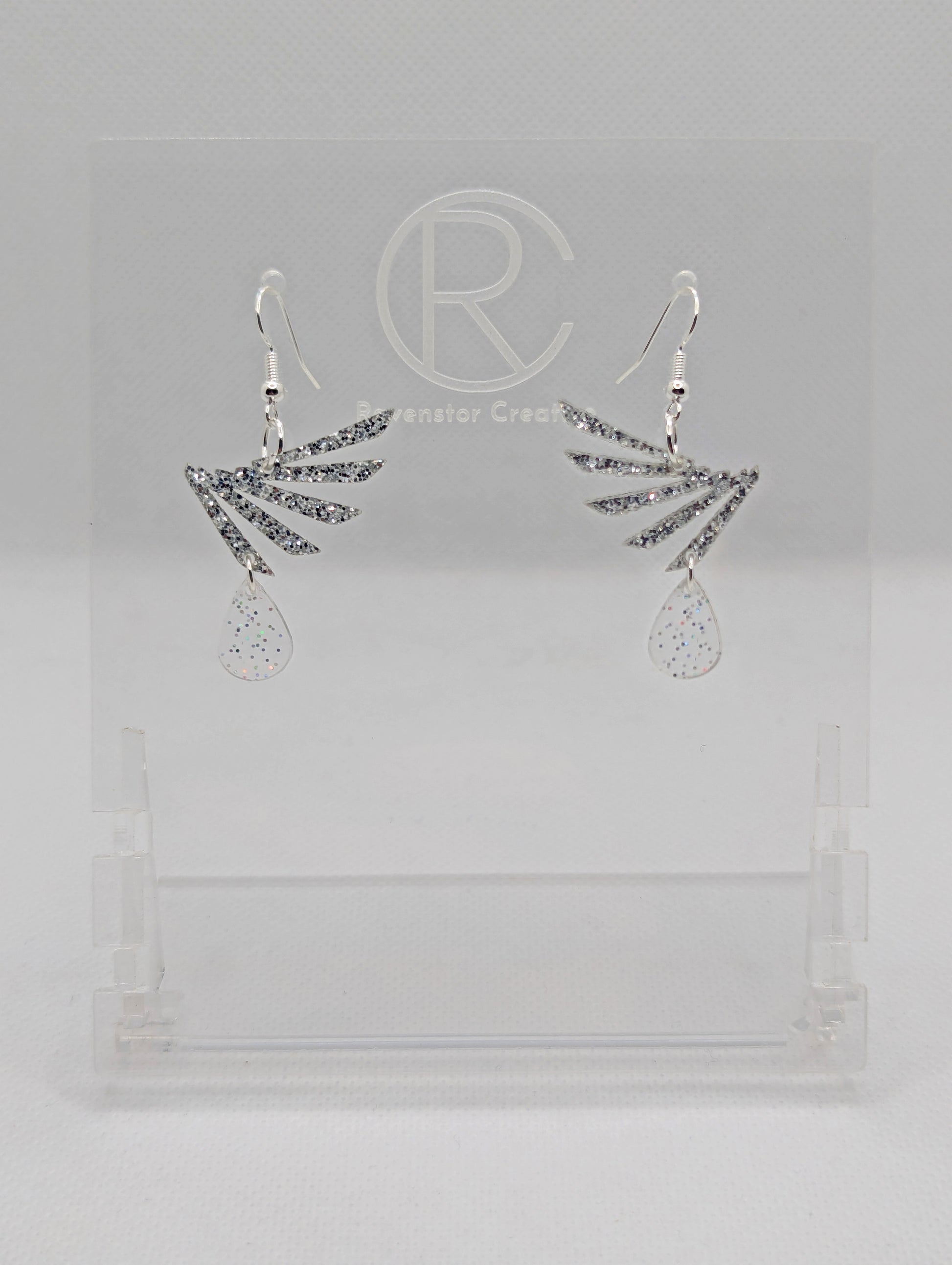 A pair of two part drop earrings with Silver ear wire hook fixings. Each one is a single wing with a teardrop balancing underneath it. The colours are Silver Glitter for the wing and Clear Sparkle for the teardrop. They are on clear stands.