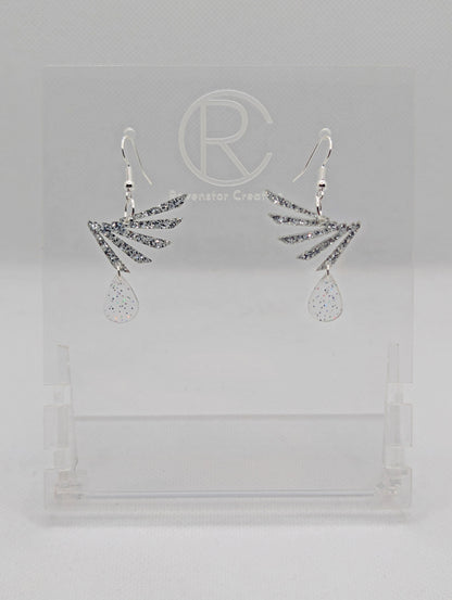 A pair of two part drop earrings with Silver ear wire hook fixings. Each one is a single wing with a teardrop balancing underneath it. The colours are Silver Glitter for the wing and Clear Sparkle for the teardrop. They are on clear stands.