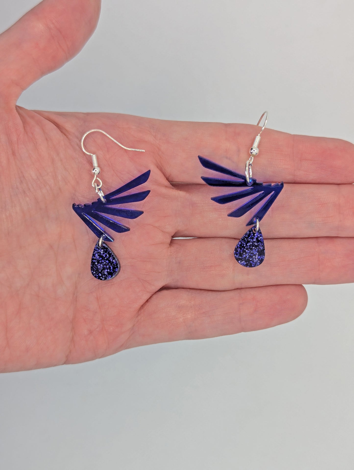 A two part drop earring with a Silver ear wire hook fixing. It is a single wing with a teardrop balancing underneath it. They are on an hand to show the size of two finger widths wide and the same for the length.