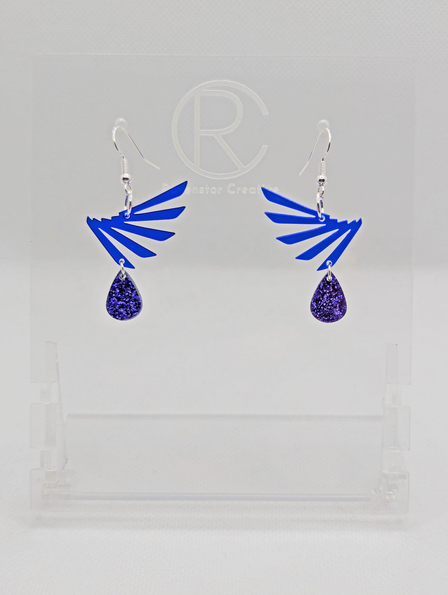 A pair of two part drop earrings with Silver ear wire hook fixings. Each one is a single wing with a teardrop balancing underneath it. The colours are Translucent Blue for the wing and Purple Glitter for the teardrop. They are on clear stands.