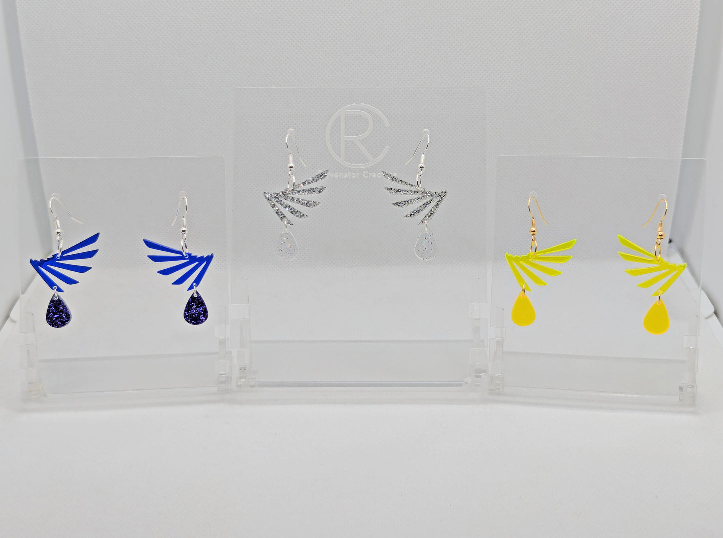 Three pairs of two part drop earrings with ear wire hook fixings. Each one is a single wing with a teardrop balancing underneath it. The colours are Translucent Blue and Purple Glitter, Silver Glitter and Clear Sparkle, Neon Yellow and Neon Orange.